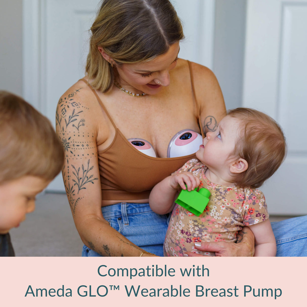 Woman wearing a brown tank top with a visible Ameda GLO wearable breast pump, holding a baby and a toddler.