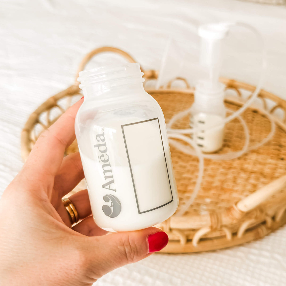 Hand holding an Ameda baby bottle partially filled with milk, with a breast pump setup on a wicker tray in the background.