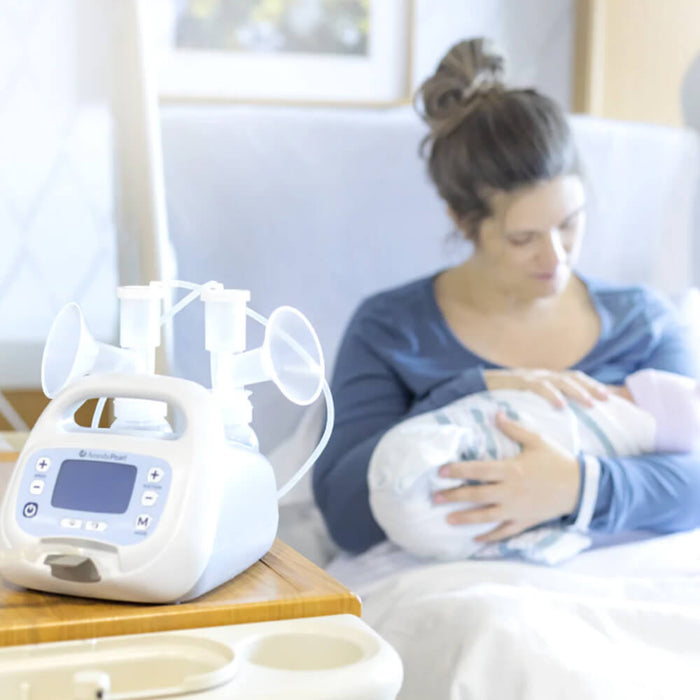 A breastfeeding mother sits in a hospital bed, gently cradling her newborn wrapped in a light blue blanket. In the foreground, a breast pump with two large funnels is placed on a bedside table, featuring a digital interface with buttons. The background is softly blurred, highlighting the mother and child. Natural light filters into the room, creating a warm and peaceful atmosphere.