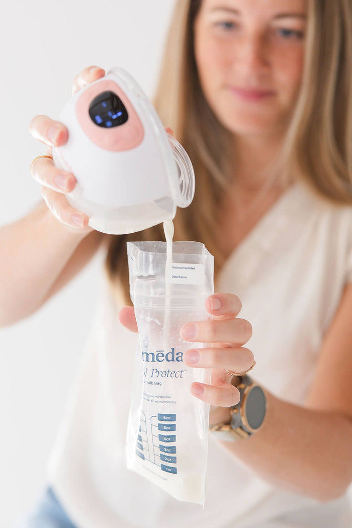 Woman pouring breast milk from an Ameda wearable breast pump into a labeled milk storage bag, demonstrating milk collection and storage process.