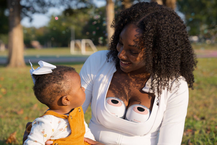 A woman sits outdoors in a park, smiling at a baby she is holding. She is using wearable breast pumps inside her top. The baby is dressed in a white shirt with floral patterns and a yellow overall and has a white bow in their hair.
