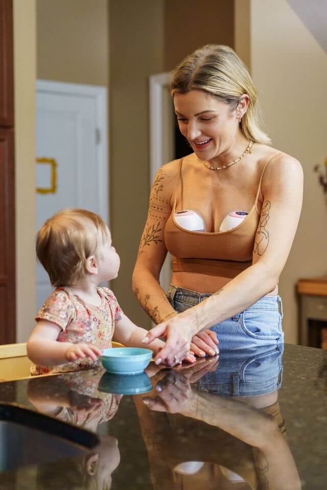 Smiling woman wearing a tan crop top and using Ameda wearable breast pumps while interacting with a young child seated at a counter with a small bowl.
