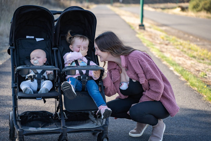 A woman kneels and smiles at two children in a double stroller on a sunny path; one child is laughing and covering their mouth, while the other has a pacifier.
