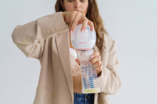 Woman in a beige blazer holds an Ameda wearable breast pump over a breast milk storage bag labeled "Pump'N Protect," transferring collected milk into the bag.