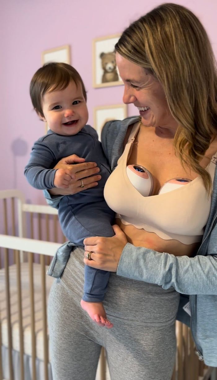 Smiling woman wearing a nursing bra with Ameda wearable breast pumps holds a happy baby in a nursery with light purple walls and framed bear artwork in the background.
