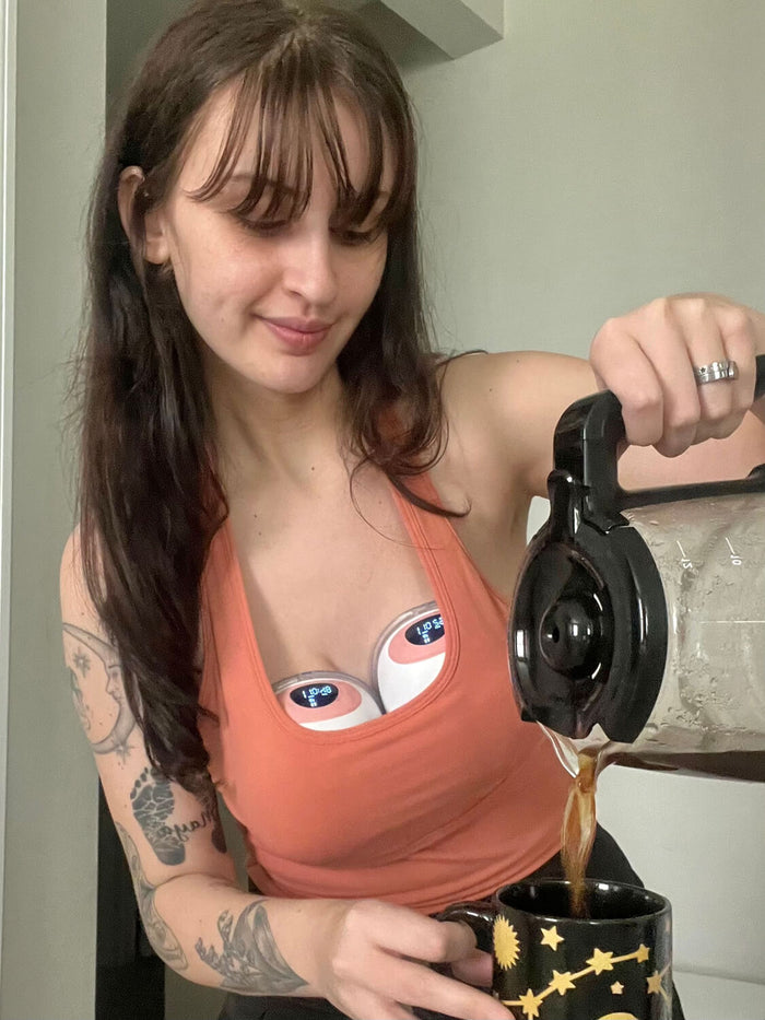 Woman wearing an orange tank top and Ameda wearable breast pumps pours coffee from a glass carafe into a black mug with a celestial design, smiling while multitasking.