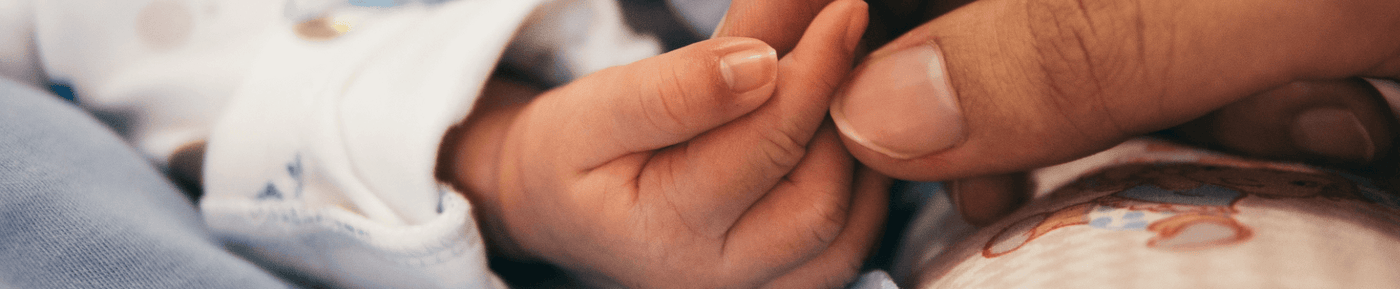 A close-up of a baby's delicate hand gently grasping an adult's finger. The baby wears a white garment with blue and light brown details, while the adult's hand is larger with a neatly trimmed nail. A patterned fabric with a teddy bear design is visible in the background, adding warmth to the tender moment.