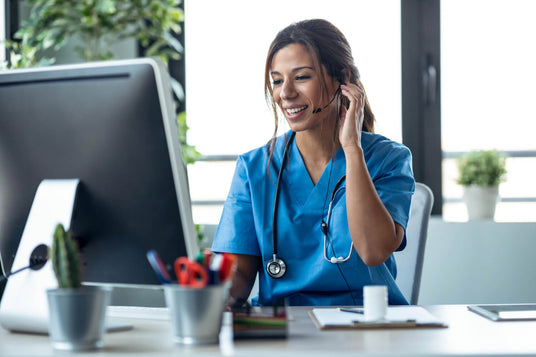 Female medical professional in blue scrubs and headset smiling while sitting at a desk with a computer.