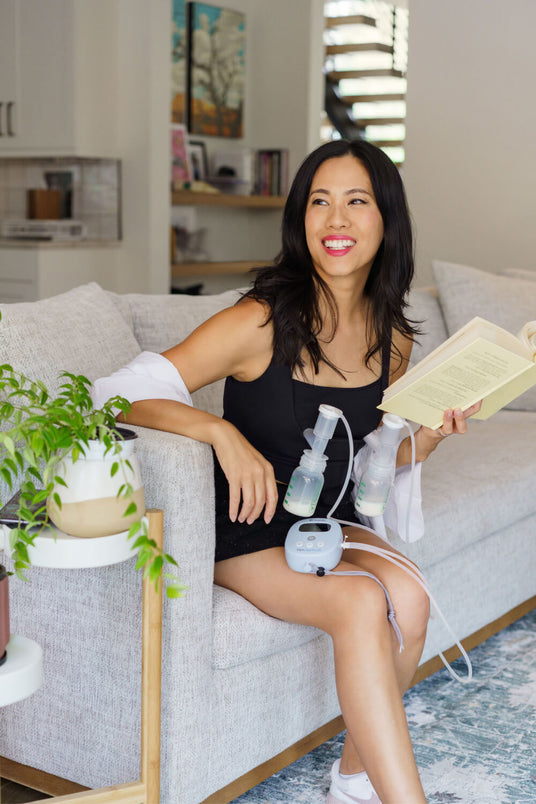 Woman sitting on a couch using a breast pump while reading a book, with a small table and potted plant nearby.