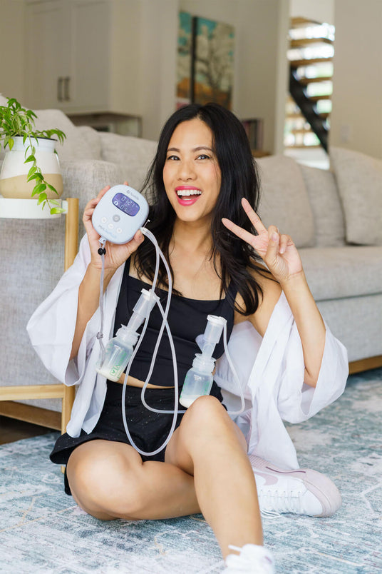 A woman sitting on the floor, smiling and holding a breast pump with attached bottles, making a peace sign with her hand.