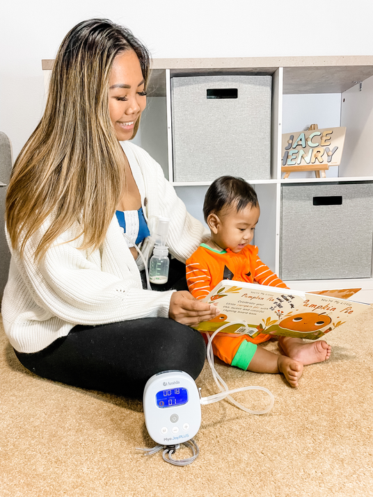 A smiling woman sits on the floor with a baby, reading a pumpkin-themed children's book. A breast pump is beside her.
