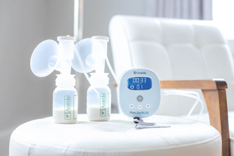 A breast pump is displayed on a round white cushion, with a sleek wooden chair visible in the background. The pump, labeled "Ameda Mya JoyPLUS," features a digital display showing a timer reading 00:33 and a pressure level at 01. Two clear plastic bottles, also labeled "Ameda," sit beside the pump, partially filled with a white liquid, likely expressed breast milk. The entire scene is illuminated with soft, natural light, creating a calm atmosphere.