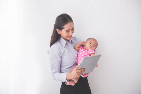 Smiling mother holding a baby in a pink polka dot outfit while looking at a tablet against a plain white background.