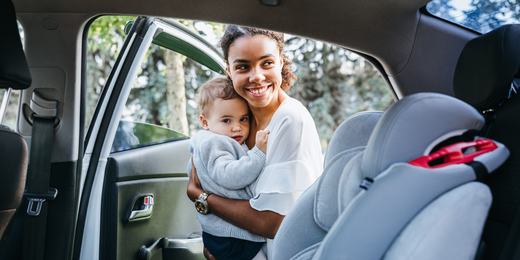 Smiling mother holding her child outside an open car door, with a car seat visible in the foreground.