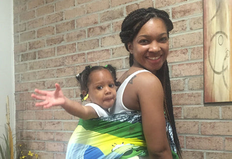 Smiling mother with braided hair carrying her child in a colorful wrap against a brick wall, while the child reaches out playfully.