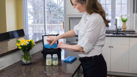 Woman in a kitchen placing an ice pack into a black breast milk cooler bag, with two breast milk storage bottles and a blue ice pack on the counter.