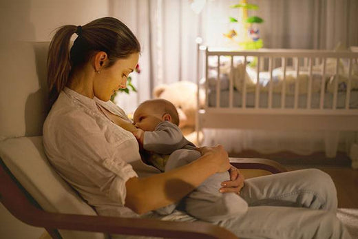 Mother sitting in a rocking chair breastfeeding her baby in a softly lit nursery with a crib in the background.