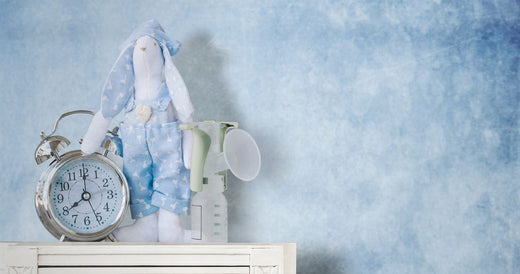 A breast pump, an alarm clock, and a stuffed bunny dressed in blue pajamas are displayed on a white shelf against a soft blue textured background.