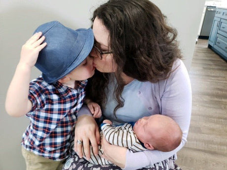 Mother holding a newborn baby while kissing her young child, who is wearing a blue hat and plaid shirt.