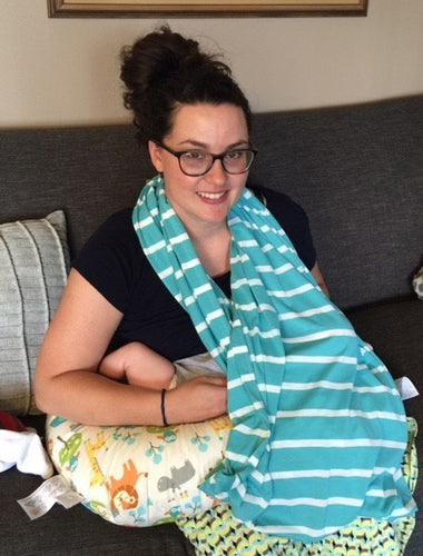 Smiling mother with glasses sitting on a couch, using a striped breastfeeding cover and a nursing pillow while holding her baby.