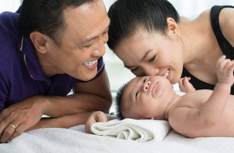 Smiling parents lying next to their baby, who is lying on a white towel, as the mother gently kisses the baby’s head.