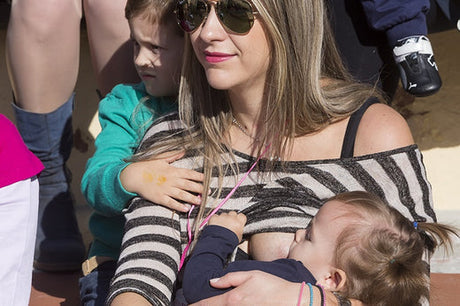 Mother wearing sunglasses and a striped top breastfeeding her baby while holding her older child, who is sitting close to her.