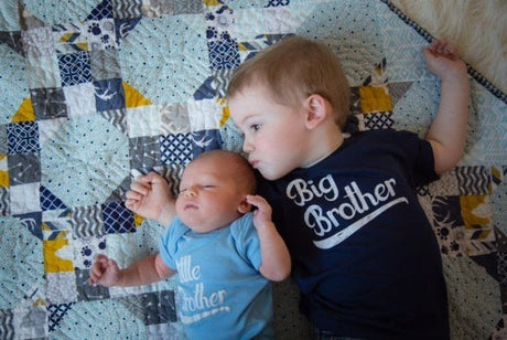 Two siblings lying on a colorful quilt; the older child wears a "Big Brother" shirt and holds the hand of the baby wearing a "Little Brother" shirt.