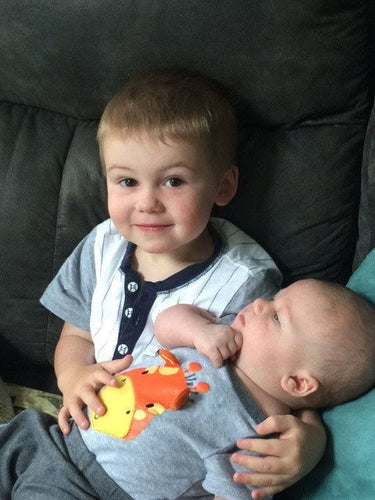 Young child sitting on a couch holding a baby sibling, both smiling and looking at the camera.