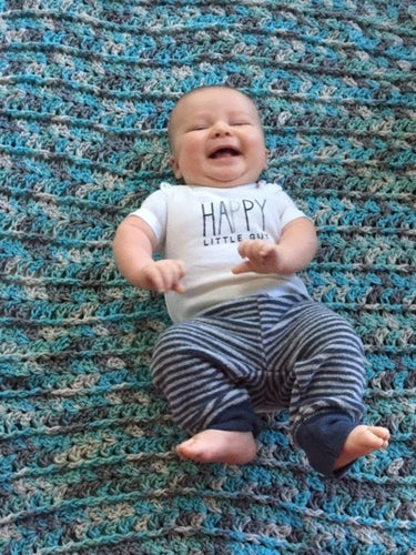 Smiling baby lying on a blue and gray crocheted blanket, wearing a "Happy Little Guy" onesie and striped pants.