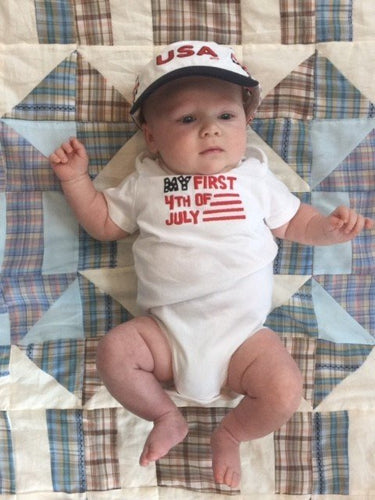 Baby lying on a quilt wearing a "First 4th of July" onesie and a USA hat, with arms and legs slightly raised.
