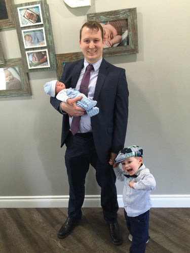 Father in a suit holding a newborn baby dressed in blue, standing next to his young child who is playfully posing with a hat indoors.