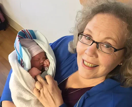 Smiling woman wearing glasses and a blue jacket holding a newborn baby swaddled in a blanket with a striped hat.