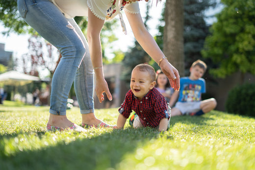 Smiling baby crawling on grass while a caregiver reaches down, with two children sitting in the background in a sunny outdoor setting.