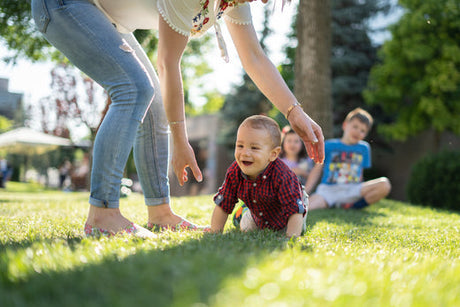 Smiling baby crawling on grass while a caregiver reaches down, with two children sitting in the background in a sunny outdoor setting.