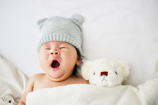 Yawning baby wearing a gray knit hat, lying in bed next to a white plush teddy bear.