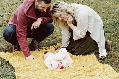 Parents kneeling on a yellow blanket in the grass, smiling down at their baby lying bundled up in a cozy outfit, during a fall outdoor activity.