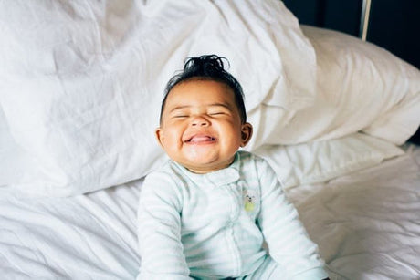 Smiling baby in light green pajamas sitting on a white bed, eyes closed and grinning widely.