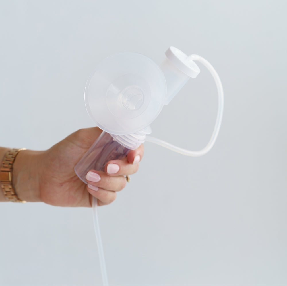 A hand holding a breast pump assembly with a transparent Ameda flange, a connected bottle, tubing, and a white valve component, shown against a light background.
