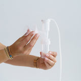 Person holding and assembling a clear Ameda breast pump with a flange, tubing, and collection bottle, demonstrating proper attachment against a light background.