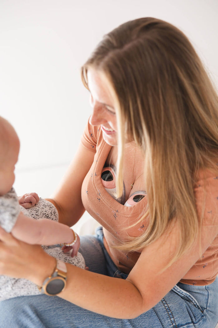 Woman wearing a peach-colored top and Ameda wearable breast pumps smiles while holding and looking at a baby, showcasing hands-free pumping and bonding time.