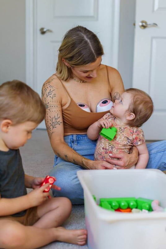 Woman wearing Ameda wearable breast pumps sits on the floor in a tan top and jeans, holding a baby and watching a young child play with toy blocks from a nearby bin.