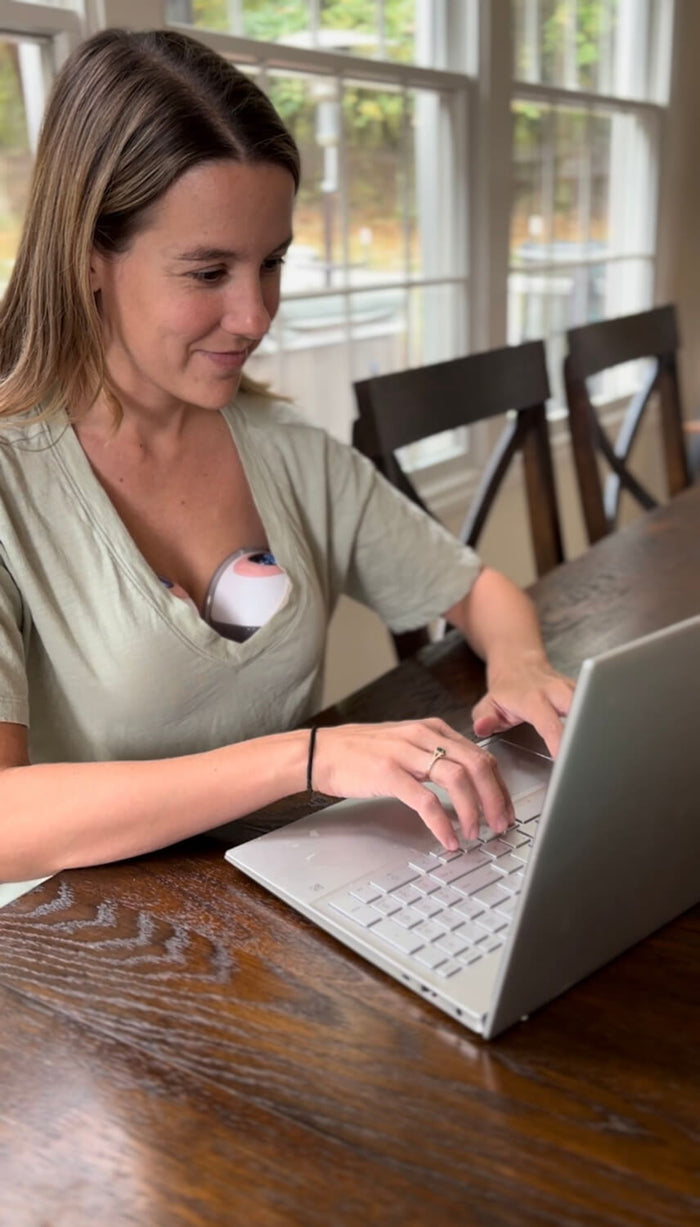 Woman wearing an Ameda wearable breast pump under a loose V-neck shirt sits at a wooden table working on a laptop, showing hands-free pumping convenience.