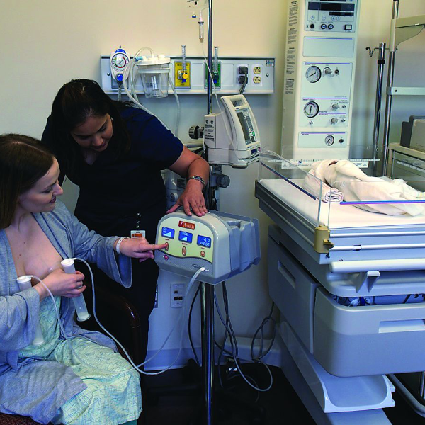 A woman in a hospital gown sits in a chair, holding two handheld devices. She looks toward a healthcare worker, who is standing beside her and pointing at a medical machine displaying digital screens and buttons. The machine is attached to various hoses and wires. Behind them is a hospital bassinet with a wrapped newborn. The environment is clinical, with medical equipment on the walls and a clean, well-lit atmosphere.
