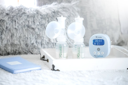 A clean, bright image of a white tray on a textured white surface holding two Ameda breast pump bottles with white lids and transparent funnels. Beside the bottles is a blue digital breast pump labeled "Mya JoyPLUS," displaying a timer of 00:33 and an expansion level of 01. A soft gray blanket in the background and a light blue notebook in the lower left corner add to the calm, soothing atmosphere.